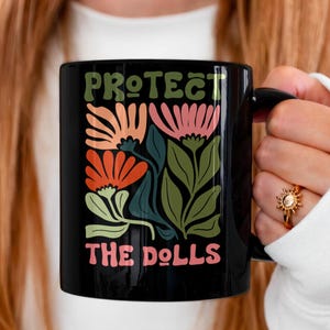 May include: Black ceramic mug with the text "PROTECT THE DOLLS" in green and pink. The mug features a floral design in orange, pink, and green. The mug has a black handle and is held by a person wearing a white sleeve.
