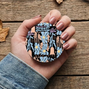May include: A round, black pop-up phone grip with a floral design in light blue, peach, and purple. The text "CALLED TO SERVE" is at the top, with "MARK 10:45" at the bottom. The grip is held in a hand.
