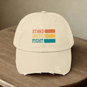 May include: A beige distressed baseball cap featuring the text STAND UNITE FIGHT printed in a stacked layout. Each word is accompanied by a horizontal rectangular block in orange, yellow, and teal, respectively, with a textured, vintage-inspired finish.