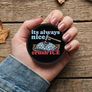 May include: A round black pin-back button featuring a graphic of a hammer crushing a bag of ice cubes. The text reads its always nice to crush-ICE in light blue and red lettering. The design is centered on a circular black background.