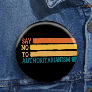 May include: A black button with a retro-style design that says "Say No To Authoritarianism" in white text with a teal, yellow, and orange striped background.