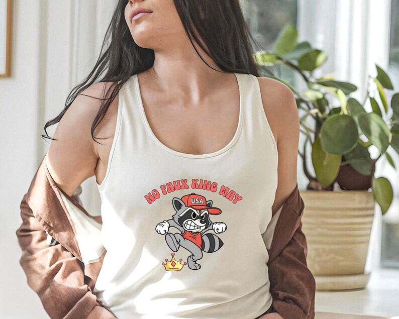May include: Cream-colored tank top featuring a cartoon raccoon wearing a red hat and shirt, with the text "NO FAUX KING MAY USA". The raccoon is kicking a golden crown. The tank top is sleeveless.