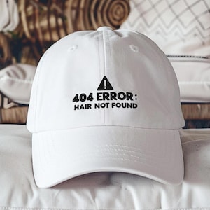 May include: A white baseball cap with a curved brim. The cap features the text "404 ERROR: HAIR NOT FOUND" in black, with a black warning symbol above the text. The cap is made of a soft material and has a classic, casual style.