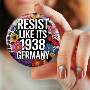 May include: A round button with the text "RESIST LIKE IT'S 1938 GERMANY" in white, set against a black background. The button is encircled by a vibrant array of colorful flowers, including pink, yellow, blue, and red blooms.