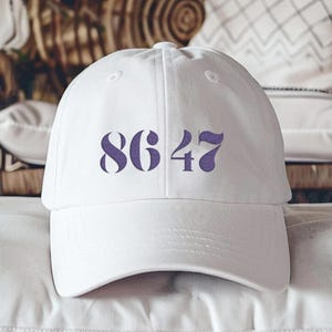 May include: White baseball cap with a purple embroidered number "8647" on the front.