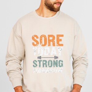 May include: A beige sweatshirt with the text "SORE TODAY STRONG TOMORROW" printed on the front. The text is in a distressed font with a green barbell graphic.