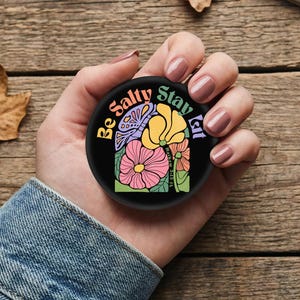May include: A black round phone grip with a colorful floral design and the text "Be Salty Stay Lit". The flowers are pink, yellow, and purple. The text is in a rainbow color scheme.
