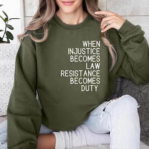 May include: An olive green crewneck sweatshirt featuring white text printed on the front that reads: WHEN INJUSTICE BECOMES LAW RESISTANCE BECOMES DUTY. The garment has long sleeves and a ribbed neckline, cuffs, and hem, providing a simple design.