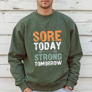 May include: Green sweatshirt with the text "SORE TODAY STRONG TOMORROW" in orange, white, and gray lettering. A small barbell graphic is above the word "STRONG".