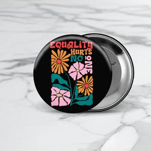 May include: A black button with a colorful floral design and the text "Equality Hurts No One".