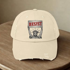 May include: A beige distressed baseball cap with a graphic of the Statue of Liberty holding a sign that says "RESIST" in red letters.