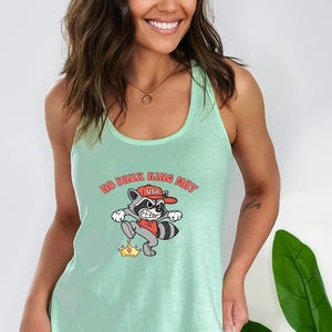 May include: A light green tank top with a cartoon raccoon graphic. The raccoon wears a red hat with "USA" on it and is stepping on a gold crown. The text "NO FAUX KING MAY" is above the raccoon.