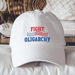 May include: White baseball cap with the words "FIGHT OLIGARCHY" in red and blue. The word "FIGHT" is above a red and white striped design with blue stars. The cap is made of a soft material.