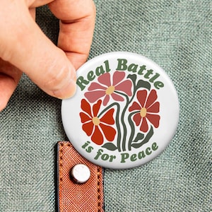 May include: A circular pin-back button with a white background featuring a graphic of three stylized flowers in shades of red and pink with dark green stems and leaves. The text "Real Battle is for Peace" is printed in dark green lettering.