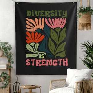May include: Black tapestry with the words "DIVERSITY is STRENGTH" in green and pink. The tapestry features colorful floral illustrations in orange, pink, and green. The tapestry is hanging on a wall with plants and furniture.