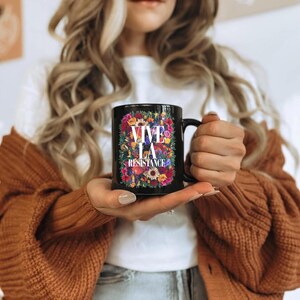 Puede incluir: Taza de cer&aacute;mica negra con la frase "VIVE LA RESISTANCE" rodeada de un dise&ntilde;o floral multicolor. La taza tiene un asa negra y la sostiene una persona que lleva una camiseta blanca y un c&aacute;rdigan marr&oacute;n.
