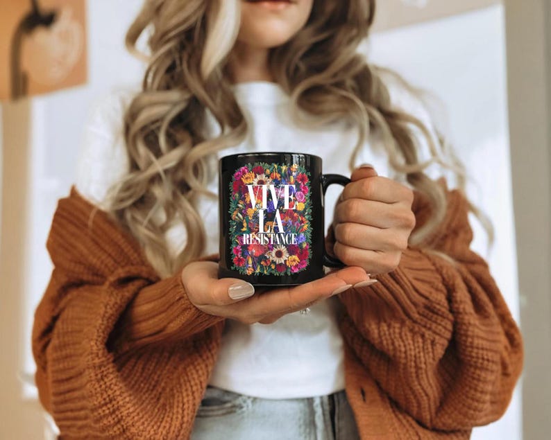 Puede incluir: Taza de cer&aacute;mica negra con la frase "VIVE LA RESISTANCE" rodeada de un dise&ntilde;o floral multicolor. La taza tiene un asa negra y la sostiene una persona que lleva una camiseta blanca y un c&aacute;rdigan marr&oacute;n.