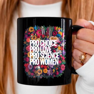 May include: A black ceramic mug featuring a vibrant, multi-colored floral arrangement surrounding the white text: PRO CHOICE, PRO LOVE, PRO SCIENCE, PRO WOMEN. The design is centered on the front of the mug, which has a sturdy handle.