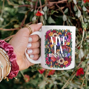 Puede incluir: Taza de cer&aacute;mica blanca con el texto "VIVE LA RESISTANCE" rodeado de un vibrante arreglo floral. La taza se sostiene sobre un fondo de follaje verde y flores rojas. La mano sostiene la taza con anillos dorados y una cinta para el pelo rosa.