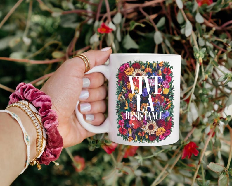 Puede incluir: Taza de cer&aacute;mica blanca con el texto "VIVE LA RESISTANCE" rodeado de un vibrante arreglo floral. La taza se sostiene sobre un fondo de follaje verde y flores rojas. La mano sostiene la taza con anillos dorados y una cinta para el pelo rosa.