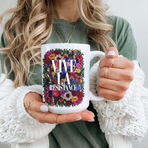 Puede incluir: Taza de cer&aacute;mica blanca con el texto "VIVE LA RESISTANCE" rodeado de un vibrante arreglo floral. La taza tiene un asa blanca y es sostenida por una persona que lleva una camisa verde y un su&eacute;ter blanco.