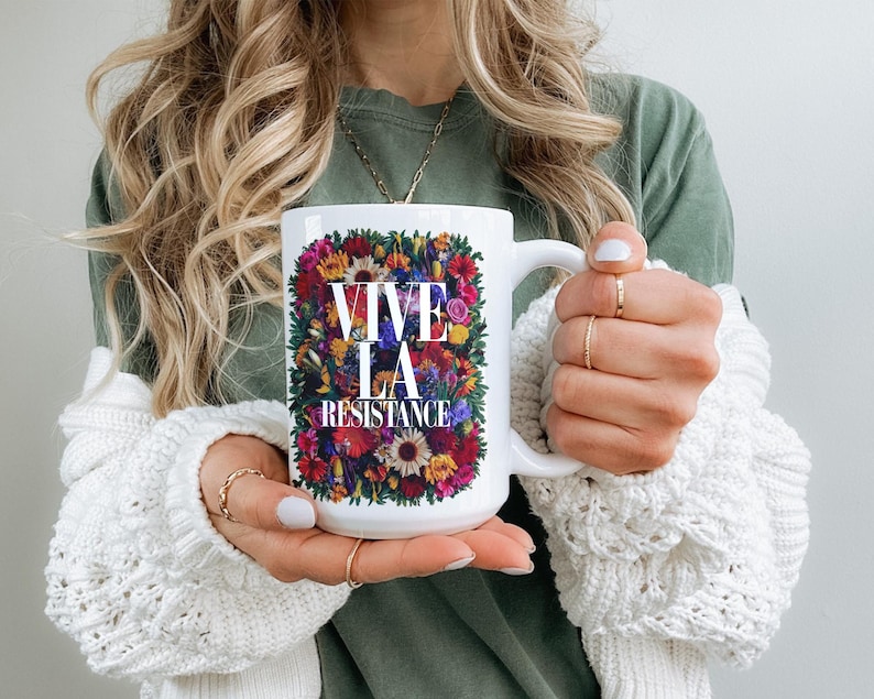 Puede incluir: Taza de cer&aacute;mica blanca con el texto "VIVE LA RESISTANCE" rodeado de un vibrante arreglo floral. La taza tiene un asa blanca y es sostenida por una persona que lleva una camisa verde y un su&eacute;ter blanco.