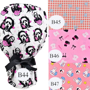 May include: A white scrub hat with a black ribbon and a pattern of pink hearts and a screaming ghost face with a phone. The hat is tied with a black ribbon. The hat is shown with three other fabric patterns: a pink and white gingham pattern, a pink pattern with black cats and white dogs, and a pink pattern with white bunnies and stars.