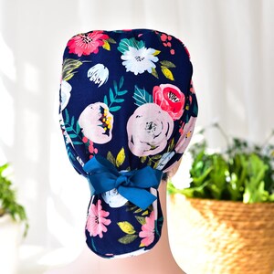 May include: A blue surgical scrub hat with a floral print featuring pink, white, and green flowers. The hat has a blue ribbon tied at the back.