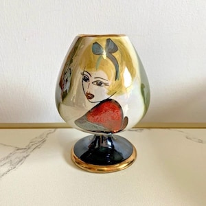 May include: A decorative, hand-painted ceramic vase with a black base and gold trim. The vase features a woman's face with blonde hair and a red dress. The vase is approximately 13 cm tall.