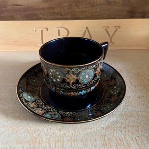May include: A black teacup and saucer set with gold and orange floral designs. The cup and saucer have a black background with gold and orange floral designs.