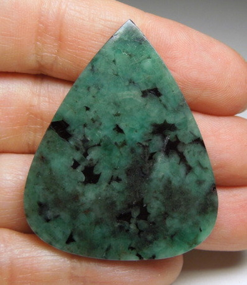 Peut inclure: Un cabochon d'&eacute;meraude verte avec des inclusions noires, en forme de larme, est pr&eacute;sent&eacute; sur une main.