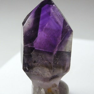 以下が含まれることがあります： クリアでファセットカットされた先端を持つ、濃い紫色のアメジストの結晶。結晶は小さな灰色の岩に載せられています。