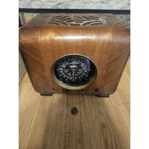 May include: A vintage wooden radio with a rounded top and a circular black dial featuring the Zenith logo and frequency markings. The radio has a decorative cutout speaker grille on top, three circular control openings, and one detached knob.
