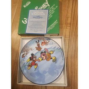 May include: A decorative plate featuring Mickey and Minnie Mouse ice skating on a frozen pond. Pluto is also depicted. The plate is in a box with a certificate of authenticity. The plate has a blue border and a winter scene.