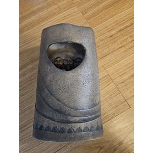 May include: A tall, gray ceramic vase with a textured surface and a large, irregular opening. Inside the opening, several small, dark faces are visible. The vase has a decorative pattern of triangles around the base.
