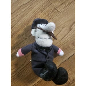 May include: A plush toy of a cartoon character with a white face, large nose, and a brown mustache. The character wears a dark blue suit, black shoes, and a matching hat. The toy has pink accents on the arms and a black bow tie.