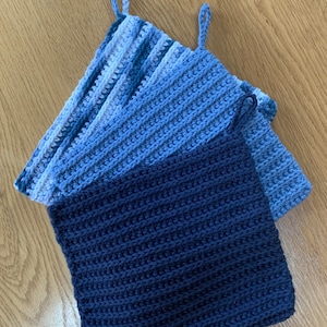May include: Three crocheted pot holders in shades of blue. The top pot holder is a gradient of light blue, dark blue, and white. The bottom pot holder is a solid dark blue. All three pot holders have a loop for hanging.