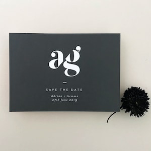 Puede incluir: Una tarjeta de guardar la fecha de color gris oscuro con texto blanco. La tarjeta presenta un monograma estilizado de las letras "ag" en blanco. El texto dice "Save the Date" seguido de "Adrian + Gemma" y "27th June 2019".
