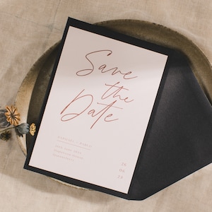 Puede incluir: Tarjeta "Save the Date" con borde negro y fondo rosa. El texto está en letra cursiva y dice "Save the Date" con los nombres "Raphael & Pablo" y la fecha "26 de junio de 2023". La ubicación se indica como "Hopetoun House, Queensferry".