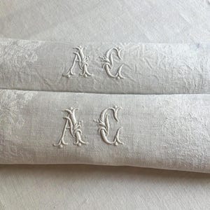 Peut inclure: Deux serviettes en lin blanc cassé pliées, ornées d'un motif floral damassé subtil et des lettres monogrammées A et C brodées en blanc au centre de chaque pièce. Le tissu présente un tissage texturé et une finition mate avec des détails brodés.