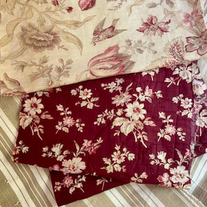 Pode incluir: Uma exibição de têxteis em camadas com um tecido superior de fundo creme e um padrão floral e de pássaros em vermelho suave, sobre um tecido bordô profundo com uma estampa floral branca densa, tudo sobre uma base listrada em bege e branco.
