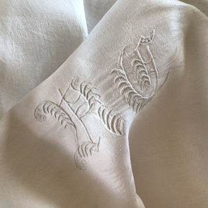 May include: White linen fabric with an embroidered design of a stylized letter H. The embroidery is done in a delicate, floral pattern.