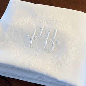 Puede incluir: Mantel de lino blanco con un patrón floral bordado y las iniciales "F.B." en el centro.