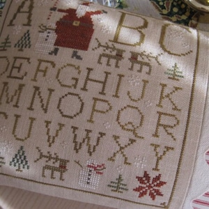 May include: A beige cross-stitch pillow with a Christmas theme. The design includes the alphabet, Santa Claus, reindeer, snowflakes, and Christmas trees. The pillow has a scalloped edge and a red and white striped fabric on the side.