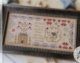 May include: Framed cross-stitch sampler with a patriotic theme. The design includes a house, American flags, and an eagle. Text reads "Where liberty dwells, there is my country." The frame is dark brown with a textured appearance.