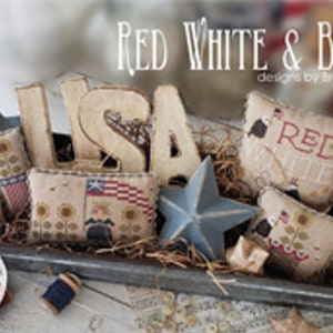 May include: A patriotic display featuring the text "RED WHITE & BLOOM" and "USA" letters. The arrangement includes star-shaped and rectangular pillows with American flag and sunflower designs, along with blue star ornaments, all within a metal tray.