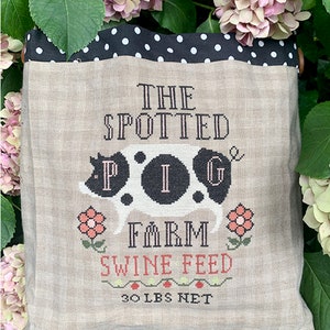 May include: A white and black spotted pig with the text "The Spotted Pig Farm Swine Feed 30 lbs Net" on a beige linen bag with a black and white polka dot trim.