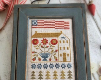 May include: A framed cross-stitch design of a house with a red, white and blue American flag above it. The house is surrounded by flowers and trees. The frame is a dark blue colour with a distressed finish. The text "THREAD WITH THY NEEDLE" is on the bottom right corner of the frame.