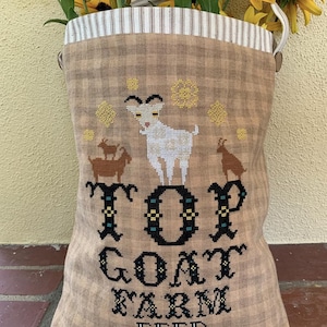 May include: A brown and tan gingham fabric bag with a white and brown striped top. The bag has a white embroidered design of three goats and the text "Top Goat Farm Feed 50lbs Net".