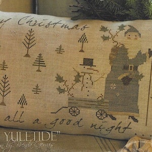 May include: A cross-stitch pillow with a beige background featuring a Santa Claus figure in blue and green, carrying a snowman on a sled. The text "Merry Christmas and to all a good night. Merry Yuletide" is stitched on the pillow.